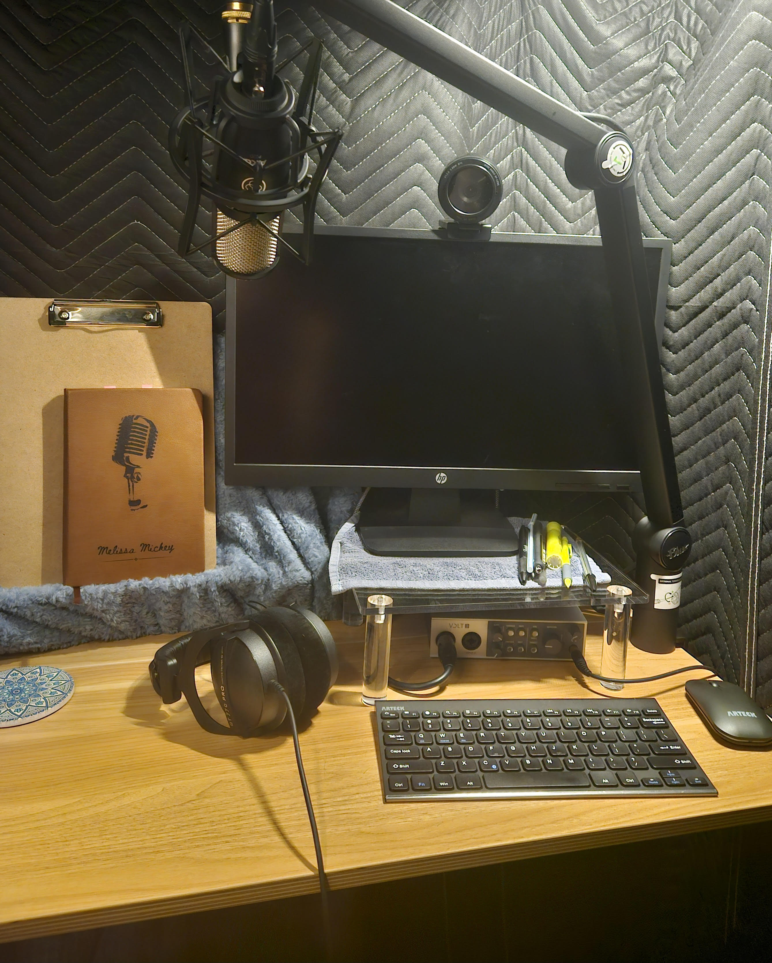 Workstation in booth, desk with keyboard, interface, paper stand with clipboard, monitor, camera, lights, microphone, and writing utensil