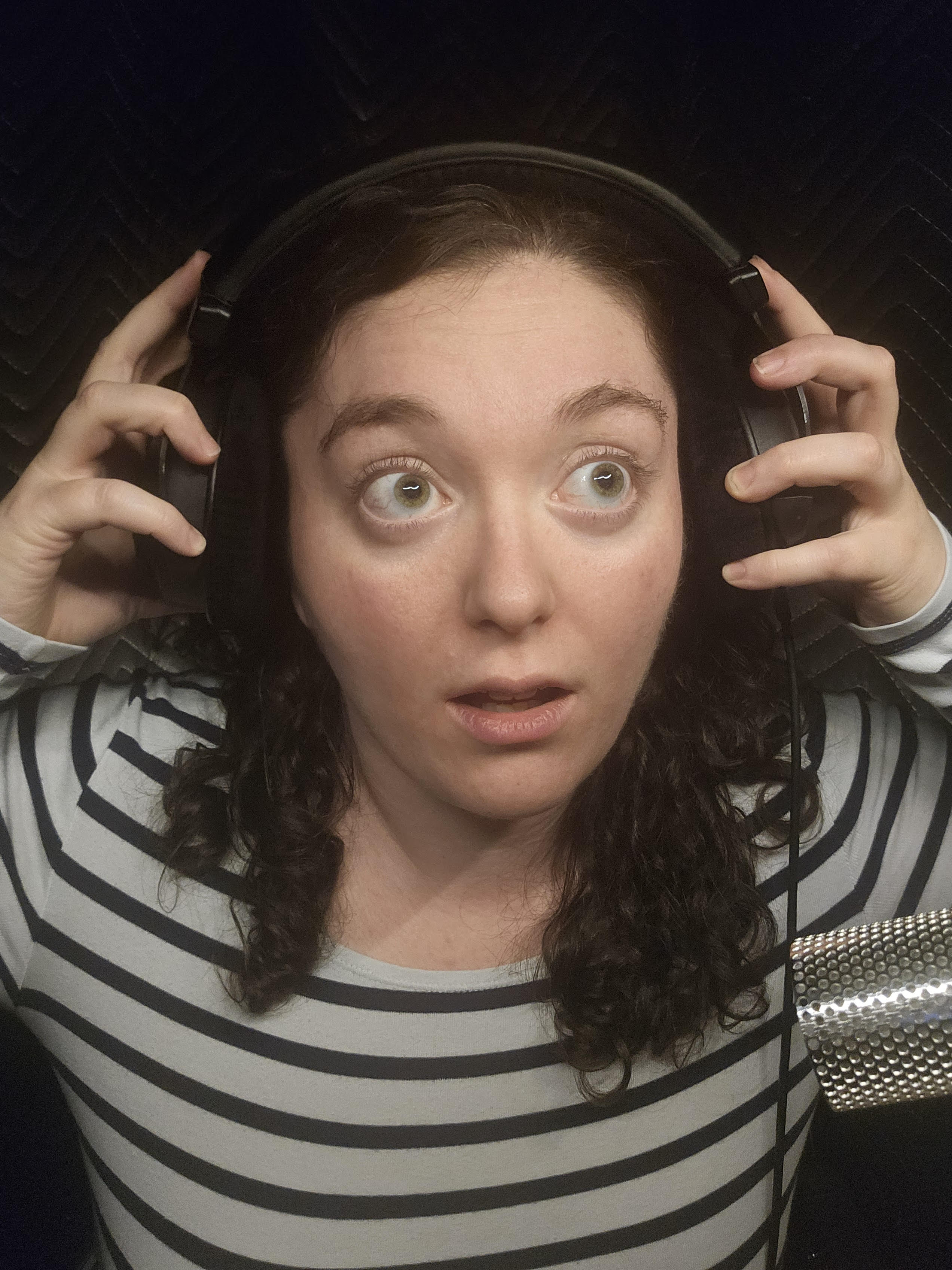 Selfie of Melissa in her booth, Headphones on, wearing glasses, standing in front of her microphone
